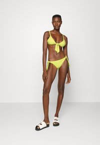 Biquíni verde neon com tecido texturizado, apresentando um top com laço à frente e parte de baixo com laço lateral. Modelo usa sandálias estilo slide brancas e pretas.