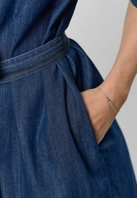 Džínové šaty s vyťahovaným pásom a plnou sukňou, s vreckom. Textúrovaná látka v hlbokej modrej farbe. Strieborný náramok viditeľný na zápästí.