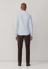 Lichtblauw, langärmig shirt met een button-down kraag, gedragen met donkerbruine broek, bekeken vanuit de achterkant.