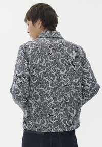 Moški v sivi in črni jakni z dolgimi rokavi s cvetličnim vzorcem, prikazan od zadaj, v temno modrih kavbojkah pred enostavno belo ozadje.