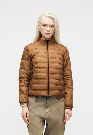 Fiatal, hosszú szőke hajú személy barna steppelt cipzáras dzsekit és világos bézs farmert visel egyszínű háttér előtt.