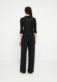 Melns caurspīdīgs trikotāžas tops ar trim ceturtdaļas piedurknēm un bārkstīm malās, kombinēts ar melnām plašām biksēm ar augstu jostasvietu un pelēkām smailām kurpēm.