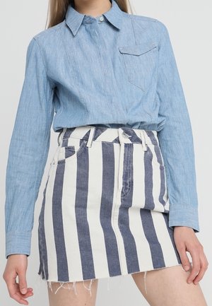 Vrouw draagt een lichtblauw shirt met lange mouwen, ingestopt in een blauw-witte, hooggetailleerde spijkerrok met verticale strepen.