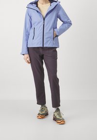 Veste imperméable bleu clair avec fermeture éclair, capuche et deux poches latérales, associée à un pantalon foncé et des baskets vertes/brunes.