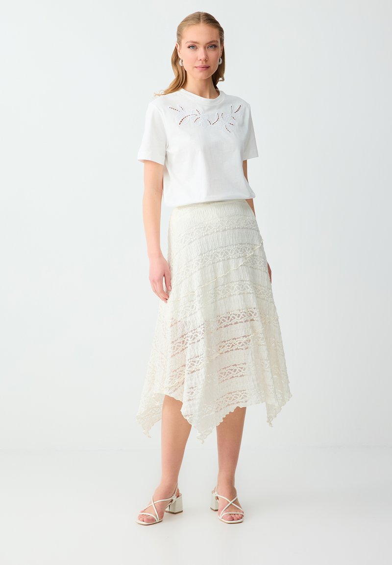 Femme portant un haut blanc à manches courtes avec des détails ajourés, associé à une jupe midi asymétrique en dentelle blanche et des talons à lanières.