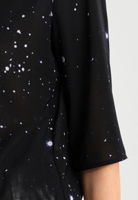 Haut noir à manches courtes avec un motif tie-dye espacé de points blancs et violets ressemblant à des étoiles sur un tissu transparent.