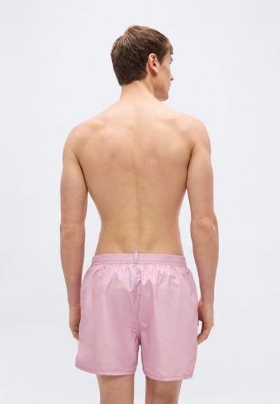 Άνδρας που στέκεται με την πλάτη προς την κάμερα, φορώντας ανοιχτό ροζ μαγιό, σε απλό ανοιχτό γκρι φόντο.