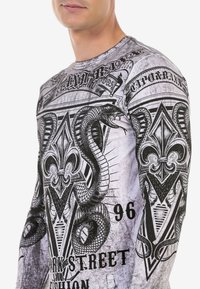 Langärmliges Shirt mit einem schwarz-weißen Grafikdesign, das eine Schlange, florale Motive und Text zeigt. Hergestellt aus leichtem Material.