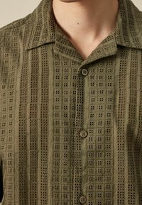 Camisa de manga corta verde oliva con tejido texturizado, que presenta un patrón de cuadrícula y recortes decorativos; cierre de botones en la parte delantera.