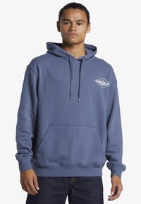 Modrá mikina s kapucňou z mäkkého materiálu, s klokaním vreckom, nastaviteľnou šnúrkou a logom na hrudi. Voľný strih a dlhé rukávy.