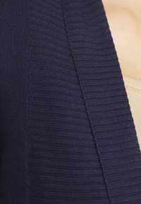 Cardigan lavorato a maglia blu navy con coste testurizzate, caratterizzato da una chiusura aperta e cuciture sottili lungo i bordi, indossato sopra un top chiaro.