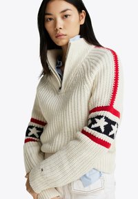 Kremowy sweter z dzianiny z pół-zamkiem, ozdobiony czerwonymi i czarnymi paskami w wzór gwiazdy na rękawach oraz teksturowanym ściągaczem na całej powierzchni.