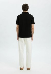 Juodas trumpas rankovėmis polo marškinėliai su tekstūruotu audiniu, derinami su laisvomis baltomis kelnėmis ir rudos spalvos šlepetėmis, matomi iš nugaros.