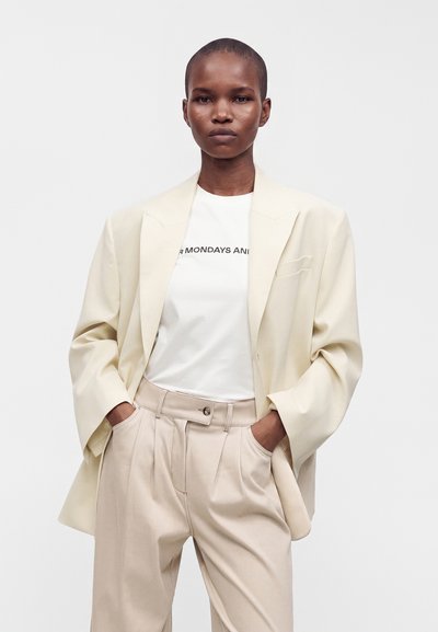 Personne portant un blazer crème oversize, un t-shirt blanc avec un texte partiel, et un pantalon plissé beige taille haute, les mains dans les poches.