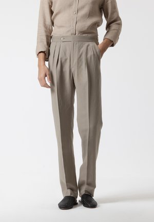 Persona con pantalones plisados beige y camisa a juego con botones, con zapatos negros tejidos tipo slip-on, de pie con una mano en el bolsillo.