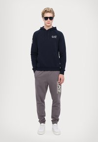 Tengerészeti kapucnis pulóver bordás mandzsettákkal, kenguruzsebbel és EA7 logóval. Szürke joggingnadrág oldalsó logóval és elasztikus mandzsettákkal. Fehér sportcipő.