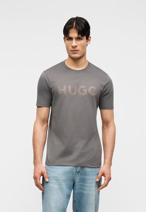 Młody mężczyzna stojący, ubrany w szarą koszulkę z nadrukiem „HUGO” na piersi oraz jasnoniebieskie jeansy na jednolitym tle.