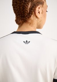 Hvit t-skjorte med svart ribbestrikket rund hals og en liten svart logo på baksiden. Stoffet ser glatt ut med en svak glans.