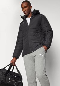 Sort pufferjakke med lynlås foran og hætte, med horisontale syninger. Grå sweatpants med logo, som holder en sort duffelbag.