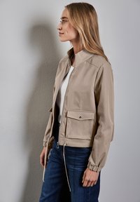 Beige bomber jacket with a zip front and elastic cuffs, featuring a chest pocket and drawstring hem. Worn over a white top and blue jeans.