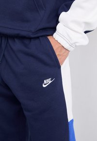 Sötétkék Nike melegítőnadrág fehér és kék panelekkel, zsinóros derékkal, puha pamut anyagból készült, és egy kis hímzett logóval a combon.