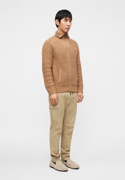 Πλεκτή καφέ ανοιχτή ζακέτα με πλαϊνές τσέπες, σε συνδυασμό με μπεζ παντελόνι cargo και μοκασίνια χωρίς κορδόνια. Απλός, υφασμάτινος σχεδιασμός.