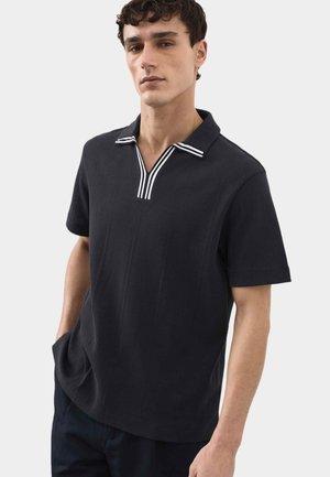 Młody mężczyzna ubrany w czarną koszulkę polo z krótkim rękawem i białym paskowanym kołnierzykiem, stoi z rękami w kieszeniach na jasnym, jednolitym tle.