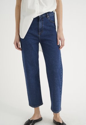 Blue denim jeans with a high waist and wide legs, featuring a front pocket, stitched detailing, and a raw hem. Pairing with a white shirt.