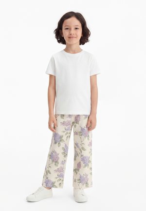 Barn med skulderlangt mørkt hår står iført en enkel hvid kortærmet skjorte, blomstrede bukser med vide ben og hvide sneakers mod en hvid baggrund.