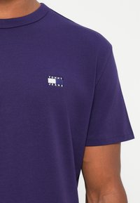T-shirt a maniche corte di colore viola in cotone con una piccola etichetta rettangolare "TOMMY JEANS" sul lato sinistro del petto. Texture liscia.