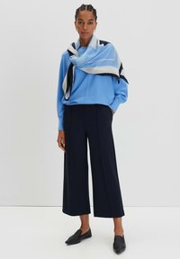 Maglione azzurro chiaro sovrapposto a una sciarpa decorata, abbinato a pantaloni ampi blu navy e scarpe slip-on nere. Trame di tessuti morbidi.