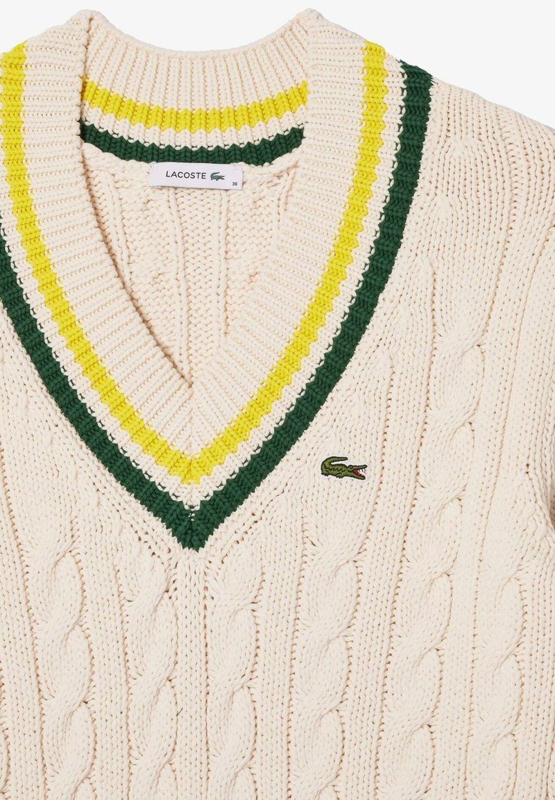 Pull en tricot à torsades crème avec un col en V rayé vert et jaune et un petit logo de crocodile vert sur la poitrine.