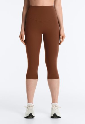 Vedere frontală a unei persoane care poartă colanți maro de lungime scurtă, cu talie înaltă, și pantofi sport albi, stând în picioare pe un fundal alb simplu.