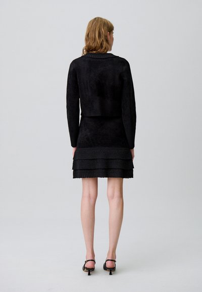 Chaqueta negra corta con un acabado texturizado, combinada con un vestido negro entallado que presenta volantes en tiers en el dobladillo. Zapatos negros de tacón bajo.