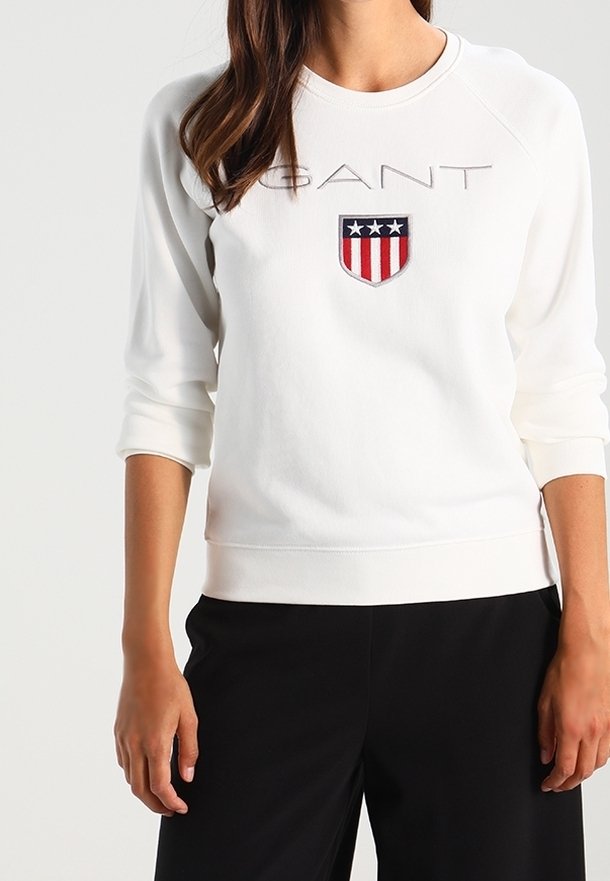 Sweat-shirt blanc à manches longues avec logo GANT et emblème de bouclier, porté avec un pantalon noir, visible du cou aux hanches.