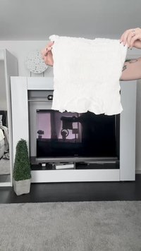 White cropped top with smocked texture, ruffled hem, and soft fabric. Held in front of a television on a modern white furniture setup.
