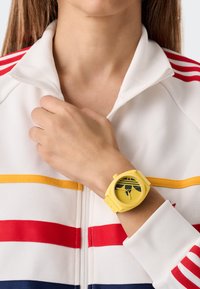 Gul silikonklocka med rund urtavla och gröna detaljer, som visas på en hand nära en vit jacka med dragkedja med röda och gula ränder.