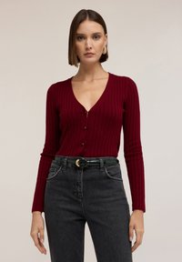 Cárdigan burdeos con canalé, escote en V y cierre de botones; combinado con jeans negros de cintura alta y un cinturón de charol verde.