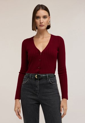 Cárdigan burdeos con canalé, escote en V y cierre de botones; combinado con jeans negros de cintura alta y un cinturón de charol verde.