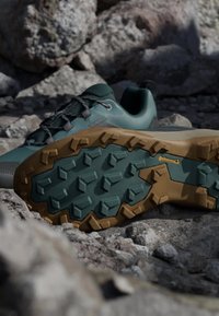 Green and brown hiking shoe with deep lug sole resting on rocky terrain, highlighting rugged grip and Continental branding on the sole.