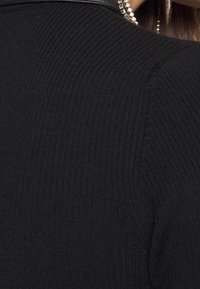 Svart ribbat tygtop med rundad halsringning och läderdetaljer vid axlarna, med en dragkedjedetalj längs halsen.