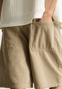 Beige bomulds shorts med to store baglommer, tekstureret stof og en løs pasform, delvist dækket af en strikket hvid top.