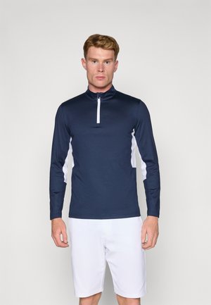 Navy long-sleeve top with white side accents, zip neck, and smooth texture. Paired with white shorts, model stands against a plain background.