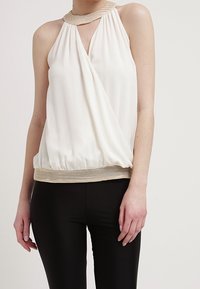 Haut blanc à col licou avec tissu plissé, présentant un décolleté en trou de serrure et une ceinture en maille dorée. Associé à un pantalon noir ajusté.