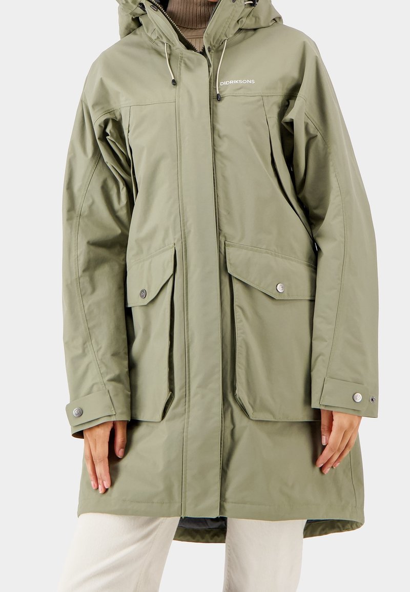 Parka imperméable verte avec capuche, dotée de deux grandes poches devant, de boutons-pression et de poignets élastiques. Texture lisse, longueur aux genoux.