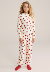 Set de pijamale din fleece cu bază crem și modele de inimioare roșii. Bluza are gât rotund, iar pantalonii au talie elastică. Șosete roșii.