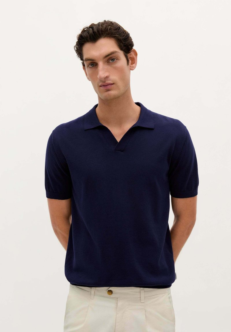 Marineblå polo shirt med en kort, lige krave, korte ærmer og en glat tekstur. Bæret sammen med lyse bukser, minimal detaljer.