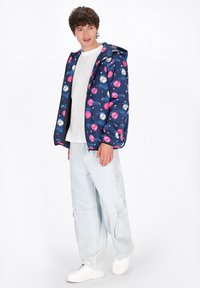 Giacca blu a fantasia con forme rosa e grigie, con zip anteriore e cappuccio, abbinata a pantaloni cargo azzurri e sneakers bianche.