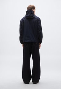 Tmavomodrý vetrovka s kapucňou, s voľným strihom a dlhými rukávmi. Kombinovaný s voľnými čiernymi nohavicami a minimálnymi detailmi.