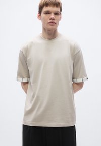 Beige Kurzarm-T-Shirt mit kontrastierenden gestreiften Bündchen. Weicher Stoff, lockere Passform, Rundhalsausschnitt und minimalistisches Design.